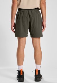 Olijfgroene shorts met een elastische tailleband, korte lengte en zijzakken. Draagt zwart schoenen en witte sokken met bedrukte tekst.