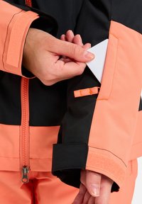 Jachetă de schi portocalie și neagră, arătând o mână care introduce un card alb într-un buzunar de pe braț. Este dotată cu mansete cu arici și detalii de cusătură în contrast.