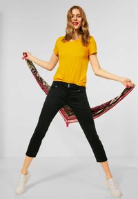 Kobieta w żółtej koszulce i czarnych spodniach trzyma w dłoni wzorzystą chustę za sobą, stojąc z rozstawionymi nogami i uśmiechając się.