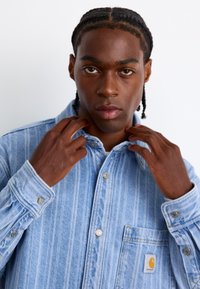 Lyseblå denim skjorte med knapper, vertikale striber, brystlomme og logo lap. Har knapmanchetter og krave. Glat tekstur.