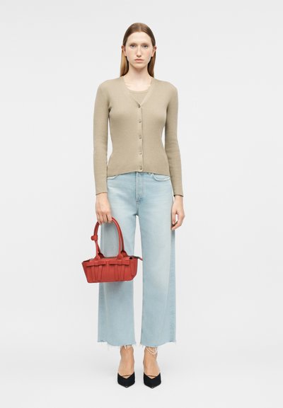 Γυναίκα που φοράει μπεζ ζακέτα με κουμπιά, ανοιχτό μπλε τζιν με φαρδιά παντελόνια και ξεφτισμένα τελειώματα, μαύρα μυτερά παπούτσια, κρατώντας μικρή κόκκινη τσάντα.