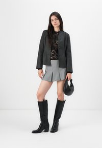 Top de encaje negro debajo de una chaqueta recortada gris, combinado con una mini falda gris con plisados y botas negras de tacón alto hasta la rodilla. Sosteniendo un bolso negro.