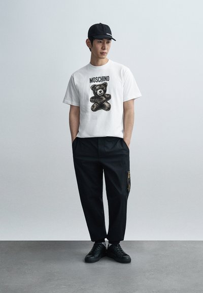 Λευκό μπλουζάκι με μαύρο γραφικό αρκούδας και το κείμενο «MOSCHINO», σε συνδυασμό με μαύρο παντελόνι με πλαϊνό φερμουάρ και μαύρα αθλητικά παπούτσια.