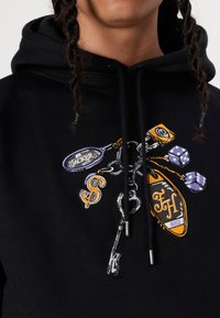 Schwarzer Hoodie mit aufwendiger Stickerei, einschließlich einer Gitarre, Spielwürfeln und Markentags in lila, orange und gelben Akzenten.