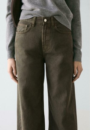 Brązowe jeansy o szerokich nogawkach, wykonane z miękkiej bawełny, z pięcioma kieszeniami, zapięciem na przód na guziki oraz gładką fakturą, połączone z siwym swetrem.