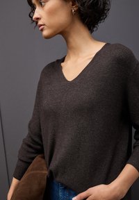 Dunkelbrauner gestrickter Pullover mit V-Ausschnitt, langen Ärmeln und lockerer Passform. Das Modell hält ein braunes Wildleder-Accessoire. Solide Textur, minimale Details.