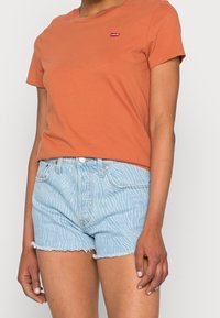Camiseta de manga corta naranja con un pequeño logo rojo de Levi's, combinada con pantalones cortos de mezclilla a rayas azul claro con bordes desflecados.