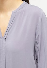 Blouse à manches longues en tissu lavande clair avec décolleté en V. Texture lisse avec un subtil éclat et des détails plissés au niveau de l'encolure.