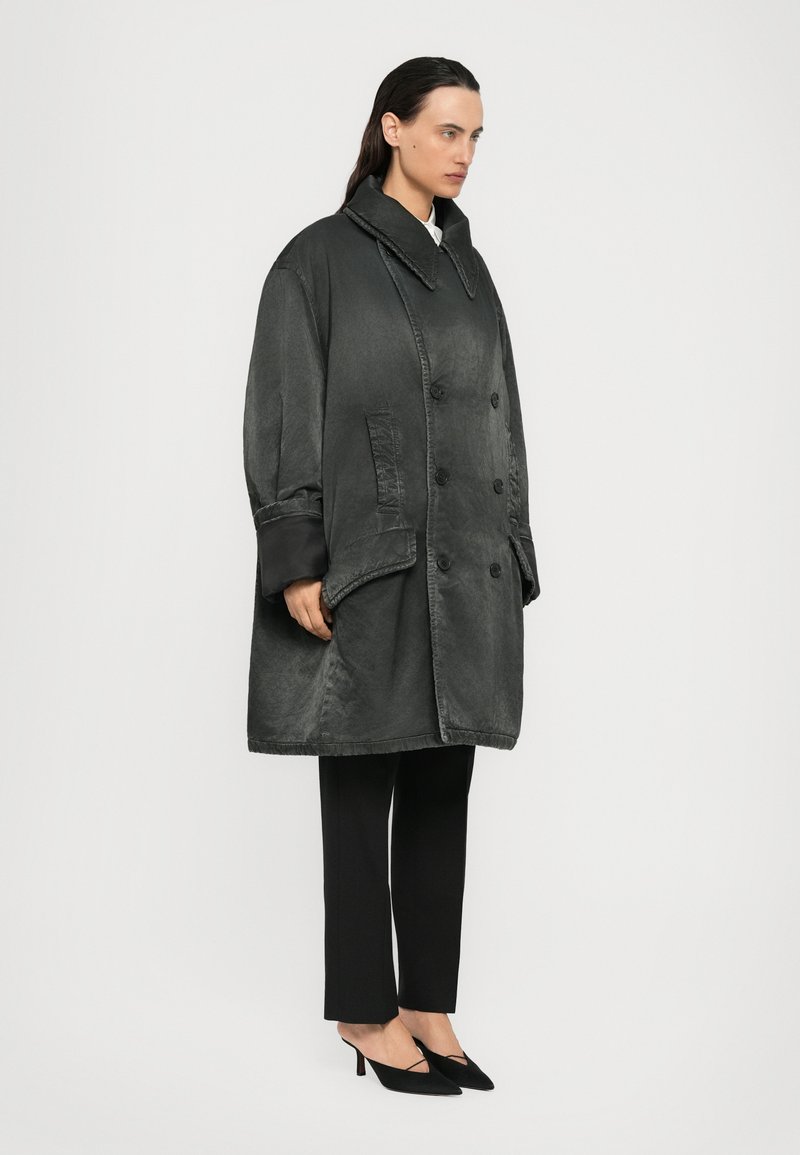 Kvinna som bär en oversized mörkgrå dubbelknäppt kappa, svarta cropped byxor och svarta pumps med spetsig tå, stående mot en enfärgad bakgrund.
