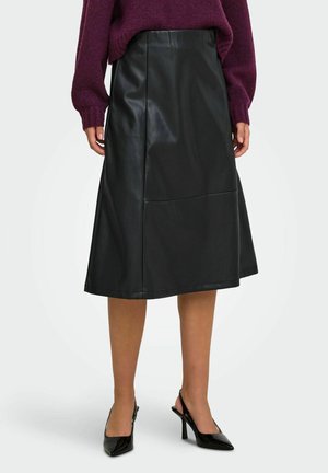 Femme portant une jupe midi en cuir noire, un pull violet et des chaussures slingback noires à talons hauts pointues, debout devant un fond blanc.