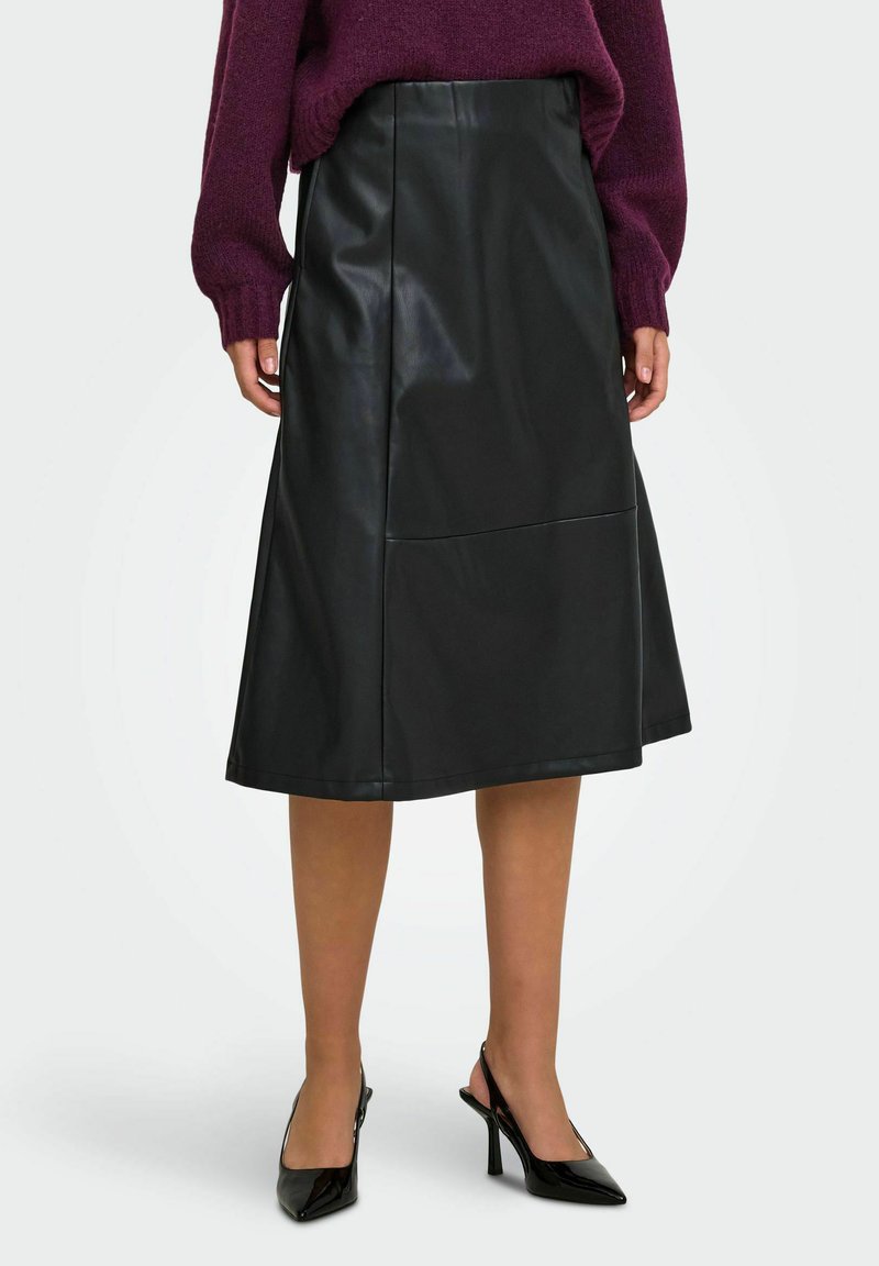Femme portant une jupe midi en cuir noire, un pull violet et des chaussures slingback noires à talons hauts pointues, debout devant un fond blanc.