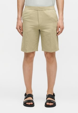 Άνδρας που φοράει μπεζ σορτς μήκους μέχρι το γόνατο με πλαϊνές τσέπες και μαύρα σανδάλια ανοιχτής μύτης, στέκεται μπροστά σε απλό λευκό φόντο.