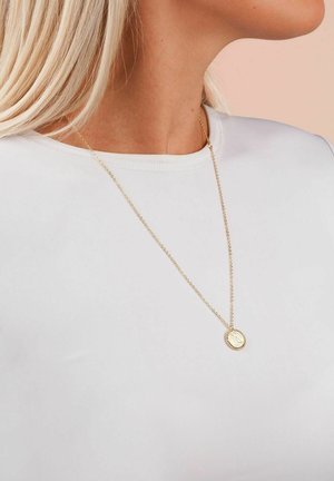 Goldene Halskette mit einem runden Anhänger, der eine strukturierte Oberfläche hat, hängend an einer filigranen Kette, getragen über einem schlichten weißen Hemd.