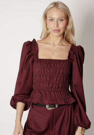 Bordowy top z bufkami, zdobiony marszczeniami i falbaniastym dekoltem. Gładka tkanina z dopasowaną talią, połączona z czarnym paskiem.