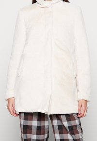 Vit plyschrock med en mjuk, fluffig textur, med hög krage och framfickor, bärs över rutiga bruna och grå byxor.