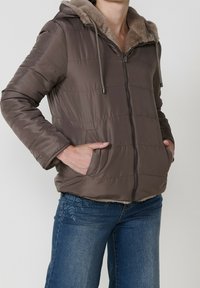 Femme portant une veste matelassée marron avec une capuche bordée de fourrure et un jean bleu, les mains dans les poches de la veste, debout devant un fond uni.