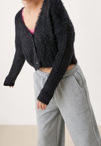 Černý huňatý kardigan s v-výstřihem a zapínáním na knoflíky, v kombinaci se světle šedými tepláky. Kardigan má strukturované pletení a dlouhé rukávy.