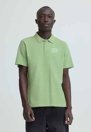 Mężczyzna w jasnozielonej koszulce polo z napisem "Palms Island" na piersi, stojący na tle jednolitego białego tła.