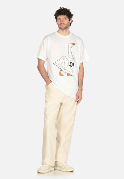 Uomo che indossa una maglietta bianca con una grafica di un'oca, una camicia oversize, pantaloni larghi color crema e sneakers beige, in piedi con una mano in tasca.