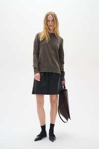 Barna pulóver, fekete bőr szoknya, fekete bokazokni és hegyes orrú balerina, sötétbarna táska a kezében. Egyszerű fehér háttér.