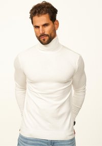 Weißer Rollkragenpullover aus gestricktem Stoff, mit langen Ärmeln und gerippten Säumen, getragen mit blauen Jeans, schlichter Hintergrund.