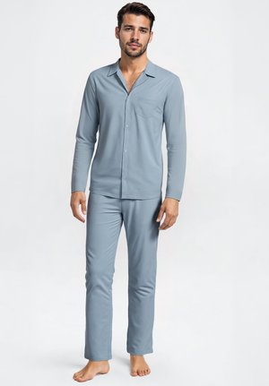 Uomo in piedi a piedi nudi che indossa una camicia a maniche lunghe celeste con bottoni e pantaloni abbinati, davanti a uno sfondo bianco uniforme.