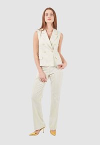 Blazer senza maniche doppiopetto crema con bottoni dorati, abbinato a pantaloni crema. Il modello indossa tacchi a punta dorati. Tessuto liscio, vestibilità sartoriale.