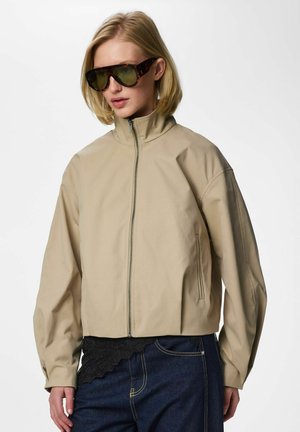 Femeie purtând o jachetă bej cu fermoar, blugi albaștri închiși și ochelari de soare cu ramă țestoasă, stând în fața unui fundal alb simplu.