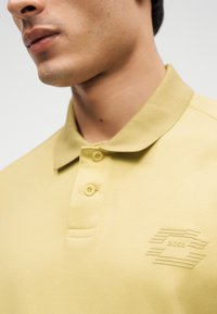 Gros plan d'une personne portant un polo jaune pâle avec deux boutons et un logo en relief subtil sur la poitrine.