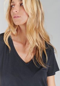 T-shirt noir à col en V fabriquée en tissu doux, avec des manches courtes et une texture lisse, portée par une personne aux cheveux longs et ondulés.