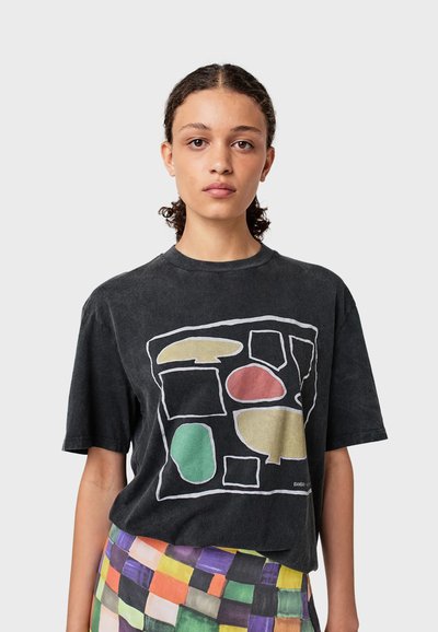 Une personne jeune avec des cheveux bouclés porte un T-shirt noir présentant des formes abstraites en beige, rouge et vert, associé à une jupe à carreaux multicolore.