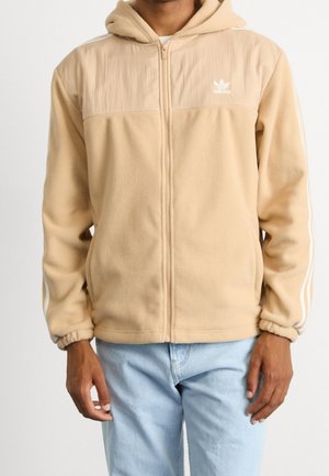 Homme portant une veste polaire beige Adidas avec capuche et bandes blanches sur les manches, associée à un jean bleu clair, debout devant un fond blanc.