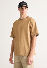 Ung mann iført en brun kortermet skjorte med stripete mansjetter og krage, beige bukser og en svart caps med "BOSS"-logo.