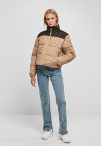 Pufferjakke med et mønstret lys brunt bryst og solid svart krage. Har en front glidelås og elastiske mansjetter. Bært med blå jeans.