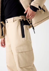 Beige Cargo-Hosen mit lockerer Passform. Sie verfügen über ein schwarzes Seitenpanel, große Taschen und Knopfverschlüsse. Das Material wirkt robust und strukturiert.