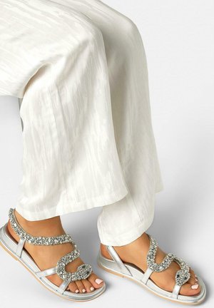 Feet wearing silver sandals with sparkling rhinestone straps paired with loose white textured pants against a plain background.