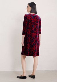 Röd sammetklänning med blommönster, trekvartsärmar, knälång skärning och rundad halsringning. Matchad med svarta platta skor.