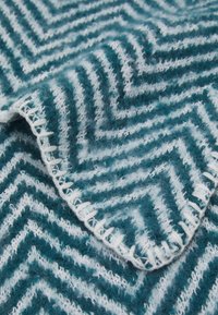 Couverture à motifs bleu sarcelle et blanc avec un design en chevrons. Texture douce avec un bord à franges et un matériau épais et moelleux.