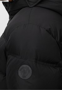 Chaqueta de plumas negra con diseño acolchado, textura suave y un parche de logo circular en el hombro izquierdo.