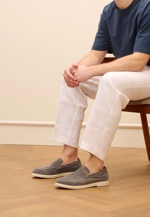 Gray suede slip-on shoes with a white rubber sole, worn with light linen pants. The design features a rounded toe and minimal stitching.