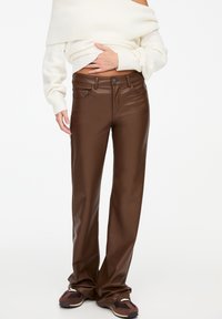 Osoba v hnědých kožených rovně střižených kalhotách, bílém svršku s odkrytémi rameny a hnědých teniskách na jednobarevném bílém pozadí.