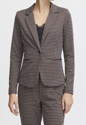 Vrouw draagt een bruin-wit geruit blazer en bijpassende broek met een zwart topje met kanten rand eronder, staand tegen een witte achtergrond.