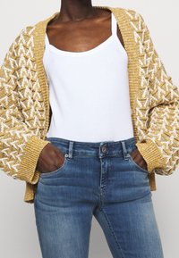 Cárdigan de punto en amarillo con un patrón geométrico blanco, de frente abierto, con puños acanalados, usado sobre una camiseta sin mangas blanca y unos jeans de mezclilla azul.