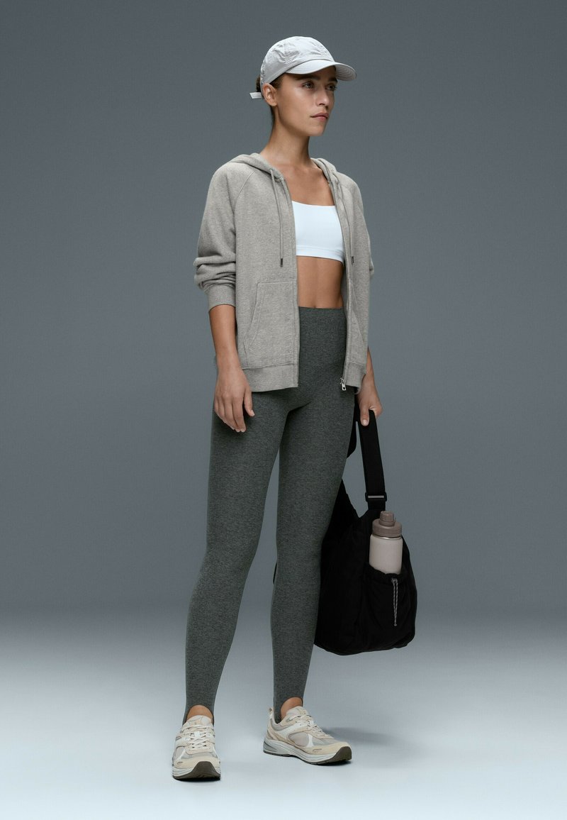 Femme en sweat à capuche gris, brassière de sport blanche, leggings gris foncé et baskets blanches, tenant un sac noir avec une bouteille d'eau, portant une casquette blanche.