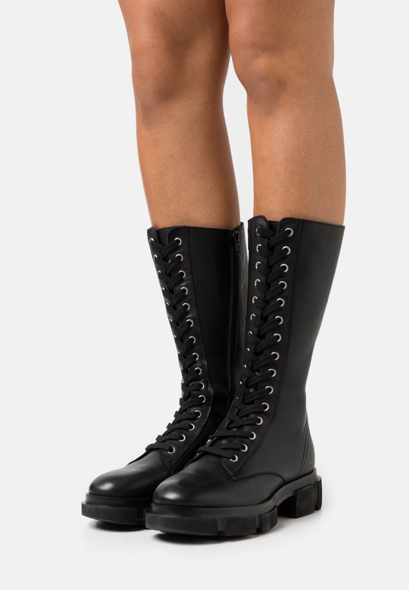 Bottes noires en cuir haute jusqu'au genou avec un laçage à l'avant, œillets en argent et une semelle en caoutchouc épaisse. Texture lisse et design structuré.