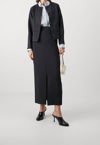 Svart cropped jacka med tryckknappar över en figursydd svart kjol med en framskjutning. Modellen håller en liten krämfärgad handväska och har på sig svarta slip-on klackskor.