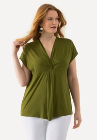 Olivově zelený top s krátkým rukávem a hlubokým výstřihem do V, se zavazováním v přední části a lehkým materiálem, doplněný bílými kalhotami.