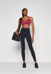 Bordó sportmelltartó, magas derekú fekete leggings perforált részletekkel a lábakon, fekete sportcipő és fekete válltáska.