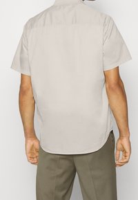 Camicia beige a maniche corte, con orlo dritto, texture liscia e dettaglio della cucitura del giromanica sul retro, abbinata a pantaloni di colore chiaro.
