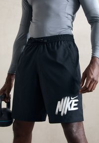 Vyras, vilkintis juodus Nike šortus ir pilką ilgomis rankovėmis marškinį, laiko juodą kettlebell'ą su mėlynu dryžiu, stovėdamas prieš paprastą foną.